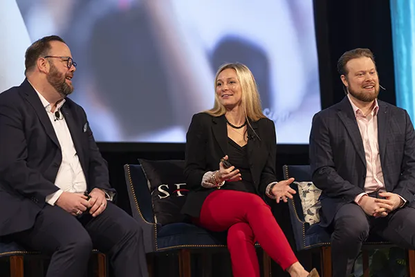 Three presenters seated on stage during a panel discussion with a large screen behind them at By Design.