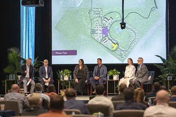 Panel of six speakers sitting on stage in front of an audience with a large projected map labeled 'Phase Two' behind them during a design session.
