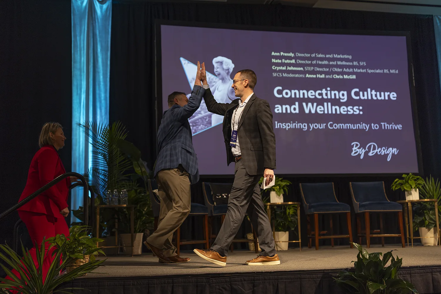 Two presenters at SFCS' By Design conference on stage giving each other a high five in front of a large screen showing the title 'Connecting Culture and Wellness: Inspiring your Community to Thrive' and speaker names.