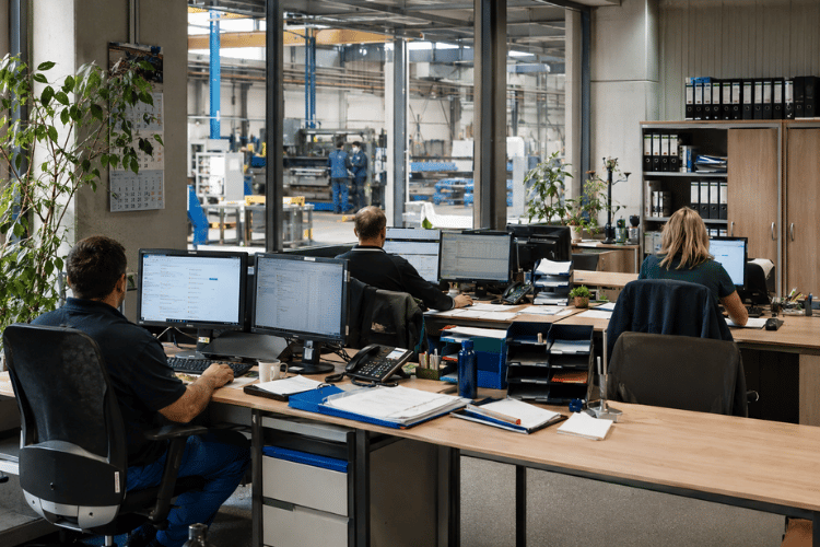 Plusieurs postes de travail dans une entreprise industrielle moderne. De grandes baies vitrées offrent une vue sur le hall de production situé derrière, où l'on aperçoit les machines et les employés à l'œuvre.
