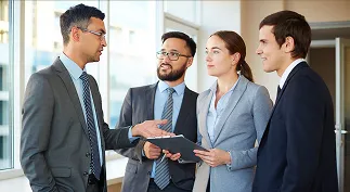 Four business professionals in suits discussing and looking at a tablet in an office setting.
