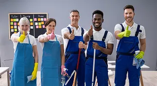 Five smiling professional cleaners in uniforms giving thumbs up, holding cleaning supplies, standing indoors with a to-do board behind them.