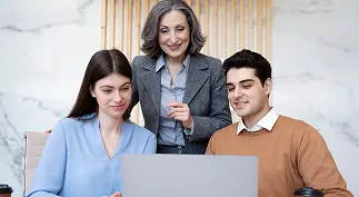 Three colleagues gathered around a laptop, with an older woman standing and two younger people seated, focused on the screen.
