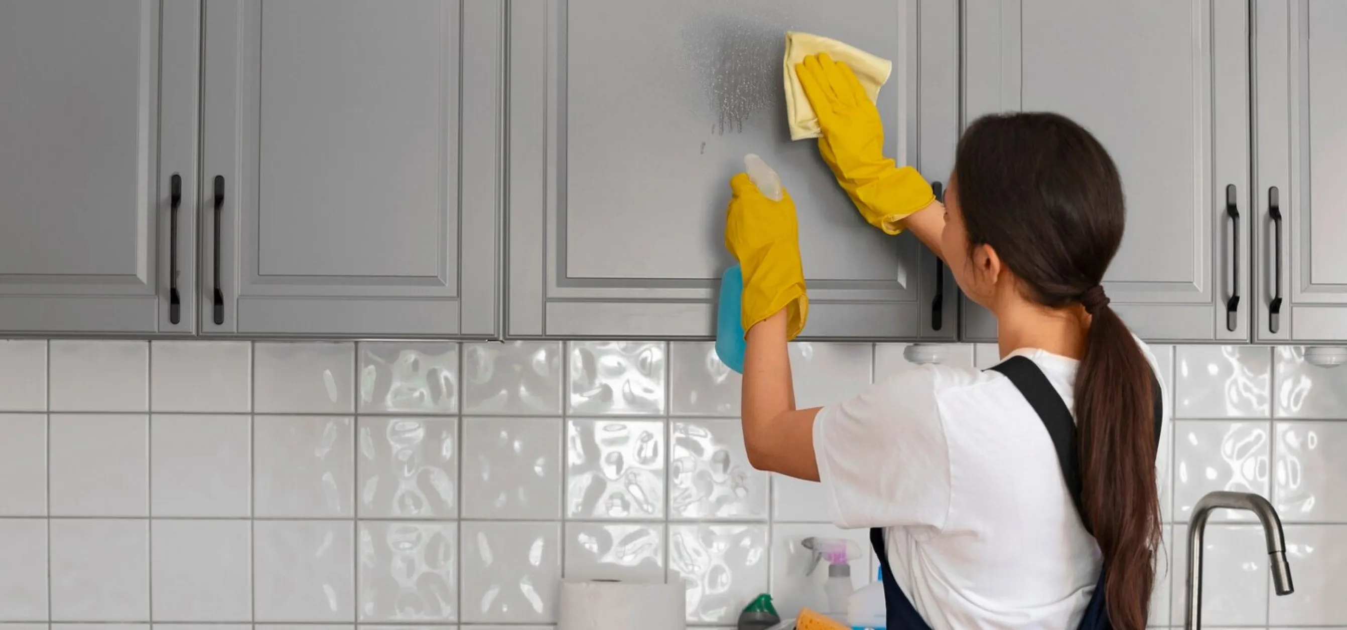 Person with long hair in a ponytail wearing yellow gloves cleaning gray kitchen cabinets with spray bottle and cloth.