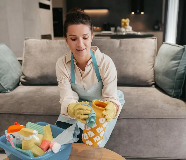 Woman wearing rubber gloves and apron cleaning a yellow vase with a cloth in a living room.
