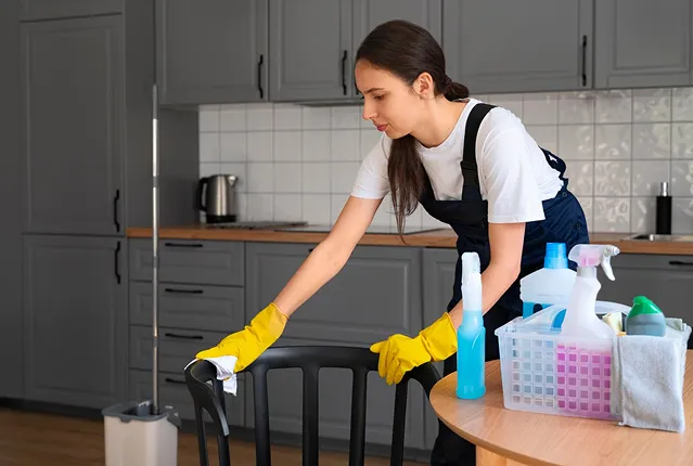 Woman wearing yellow gloves cleaning a black chair in a modern kitchen with cleaning supplies on a nearby table.