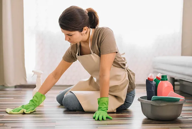 Woman wearing green gloves and apron kneeling while cleaning a hardwood floor with a cloth, next to a basin with cleaning products.