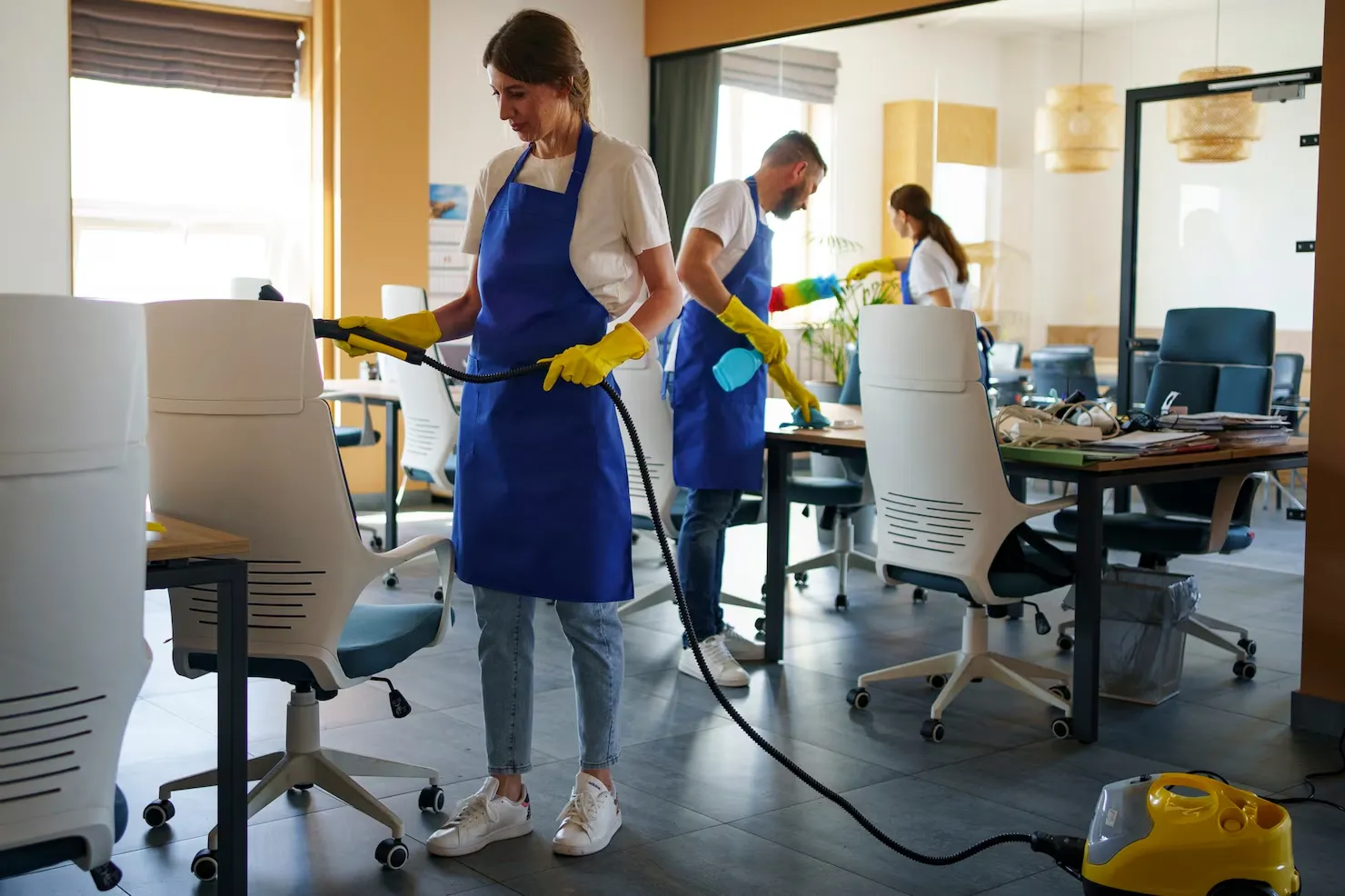 Three janitors wearing blue aprons and yellow gloves cleaning an office space with white chairs and desks.