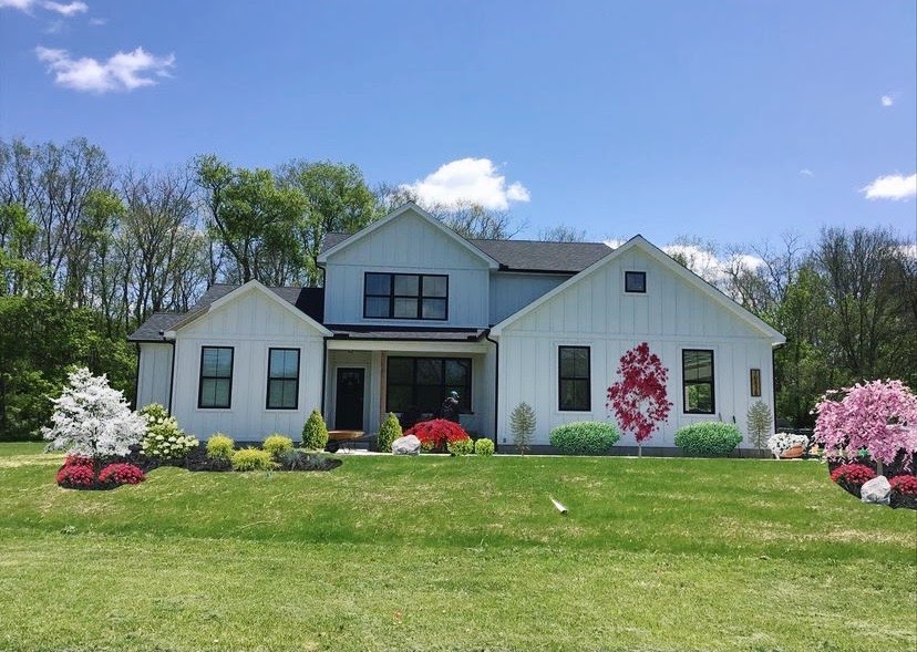 Modern white farmhouse with black trim and colorful spring landscaping featuring red, pink, and white flowering shrubs.