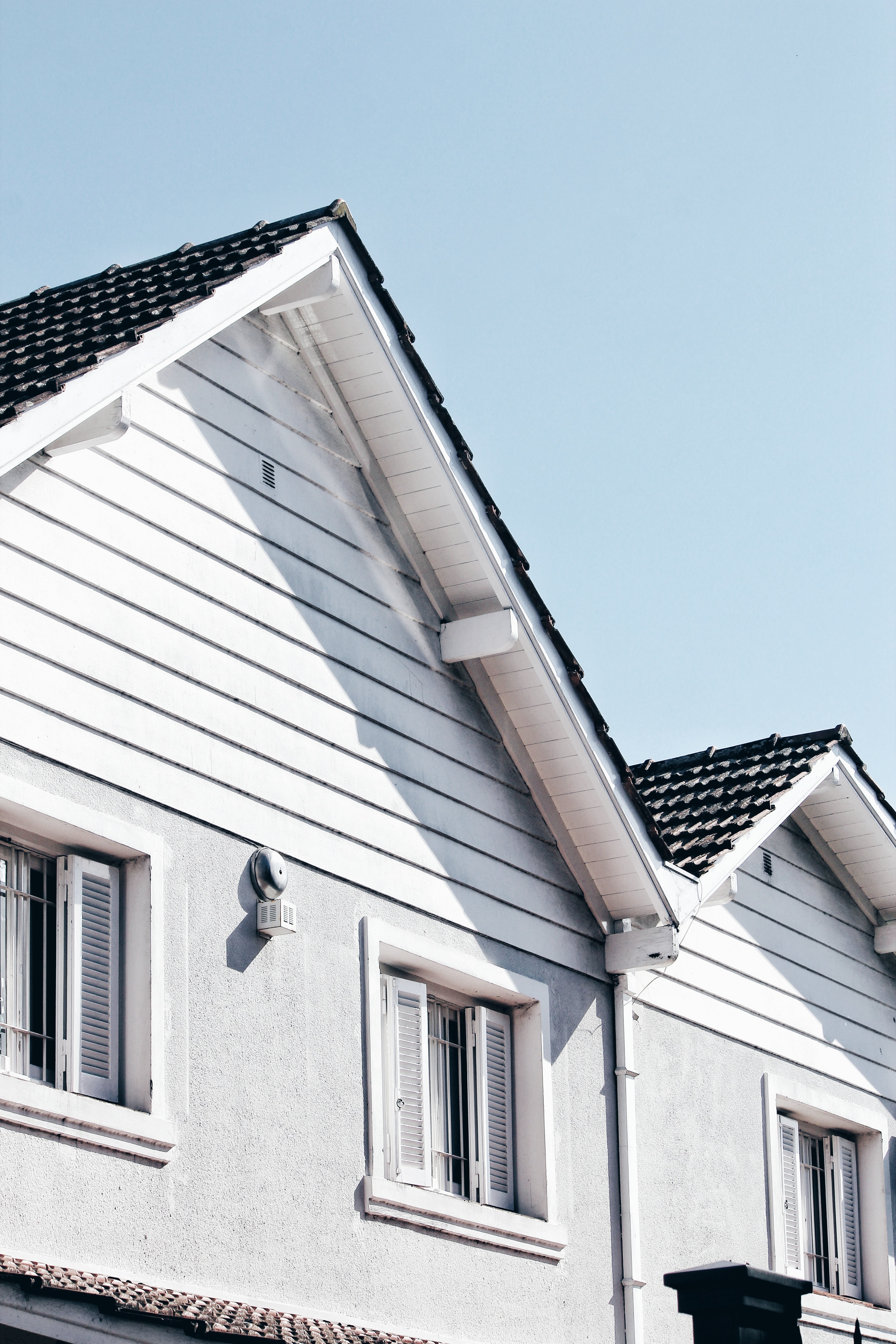 Close-up of a modern white house exterior with clean siding, windows, and dark roof tiles under clear blue sky, showcasing spring home improvement appeal.