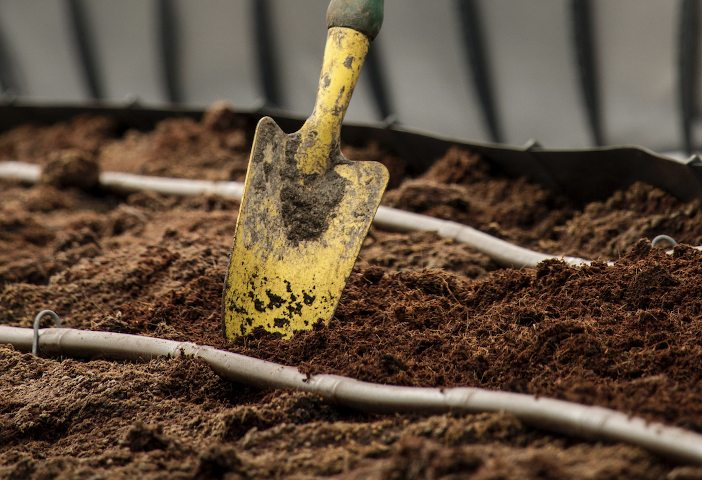 Close-up of a yellow garden trowel in nutrient-rich soil with drip irrigation lines, illustrating soil preparation for a healthy and thriving landscape.
