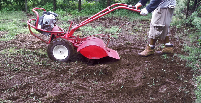 A person using a red rototiller to prepare garden soil, breaking up compacted dirt to improve aeration and create a healthy foundation for new landscaping.