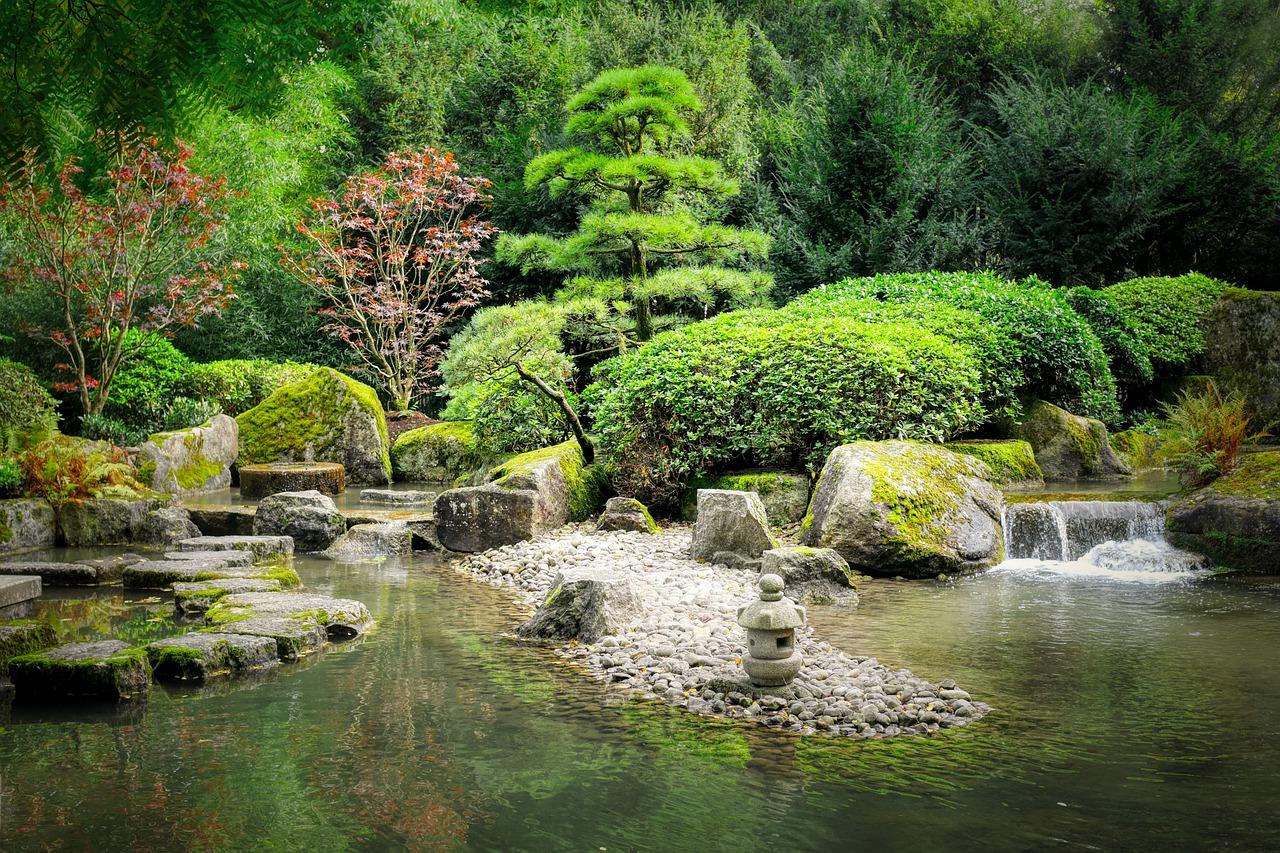 Tranquil Japanese-style garden with moss-covered rocks, lush green shrubs, stepping stones, and a small waterfall flowing into a peaceful pond.