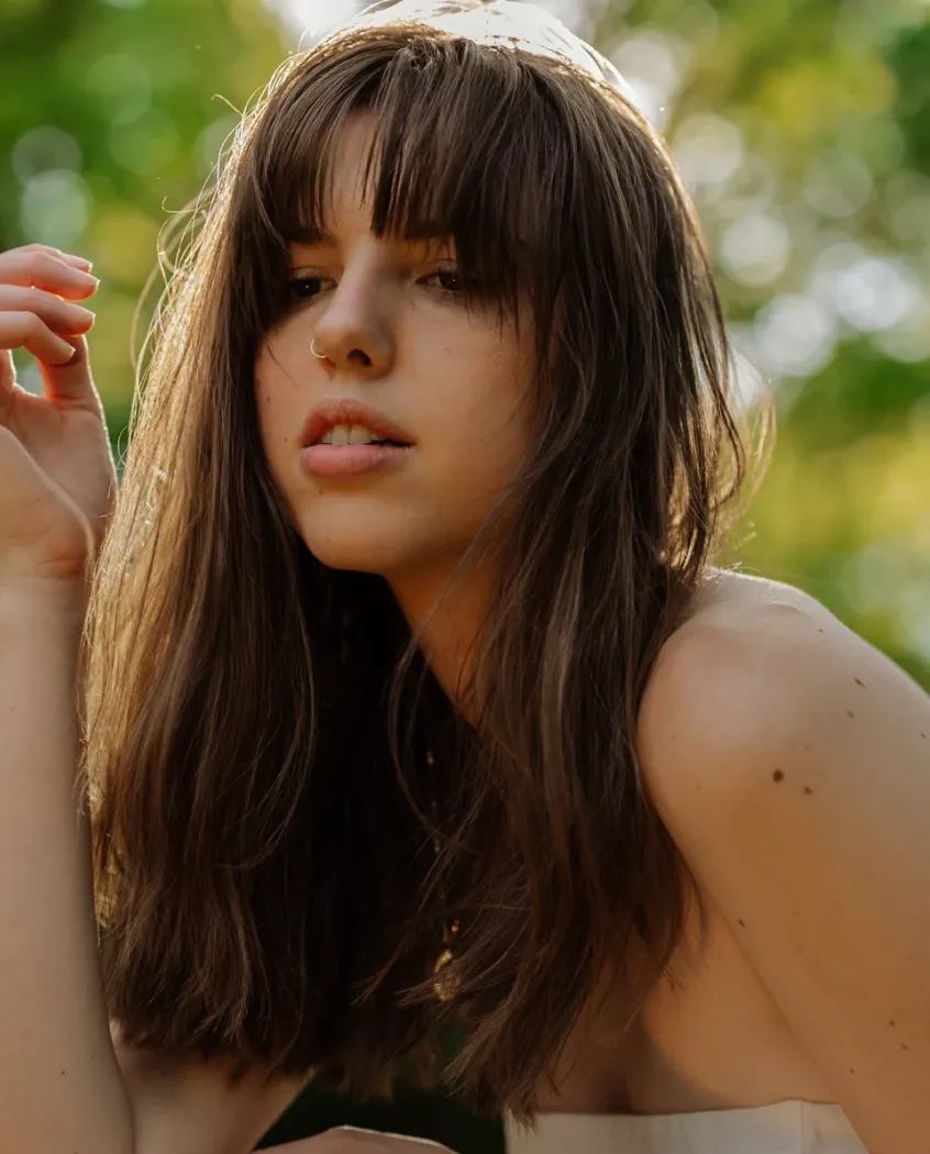Young woman wearing a knitted hat with a pom-pom and a blue jacket, outdoors near rocky terrain at dusk.