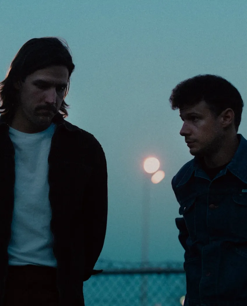 Young woman wearing a knitted hat with a pom-pom and a blue jacket, outdoors near rocky terrain at dusk.