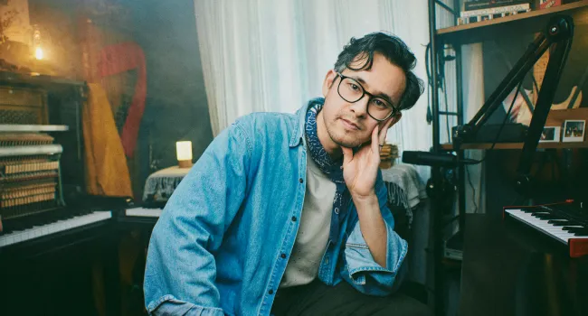 Young man with glasses and scarf leaning his head on hand seated in a cozy room with musical instruments.