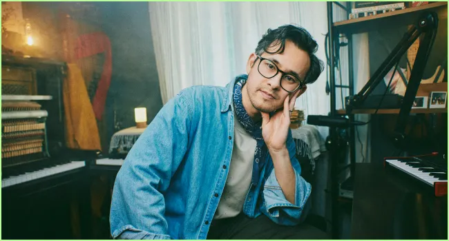Young man with glasses and scarf leaning his head on hand seated in a cozy room with musical instruments.