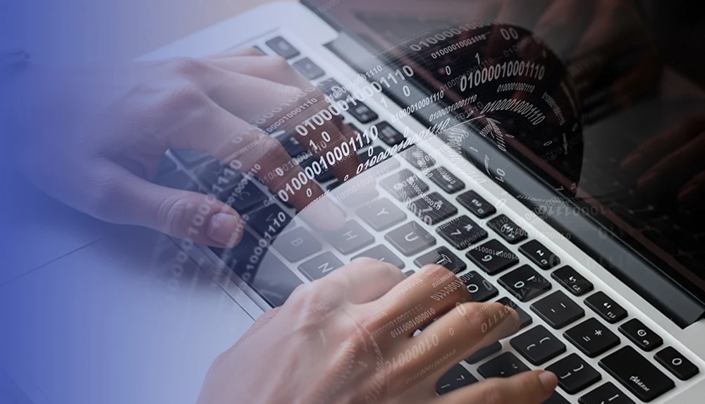 Close-up of a laptop screen and the hands of a person writing on a notebook. In the foreground a link chain with digital binary codes on it.