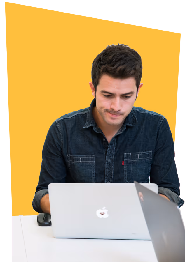 Young man in a dark denim shirt focused on his laptop at a white desk with a yellow background.