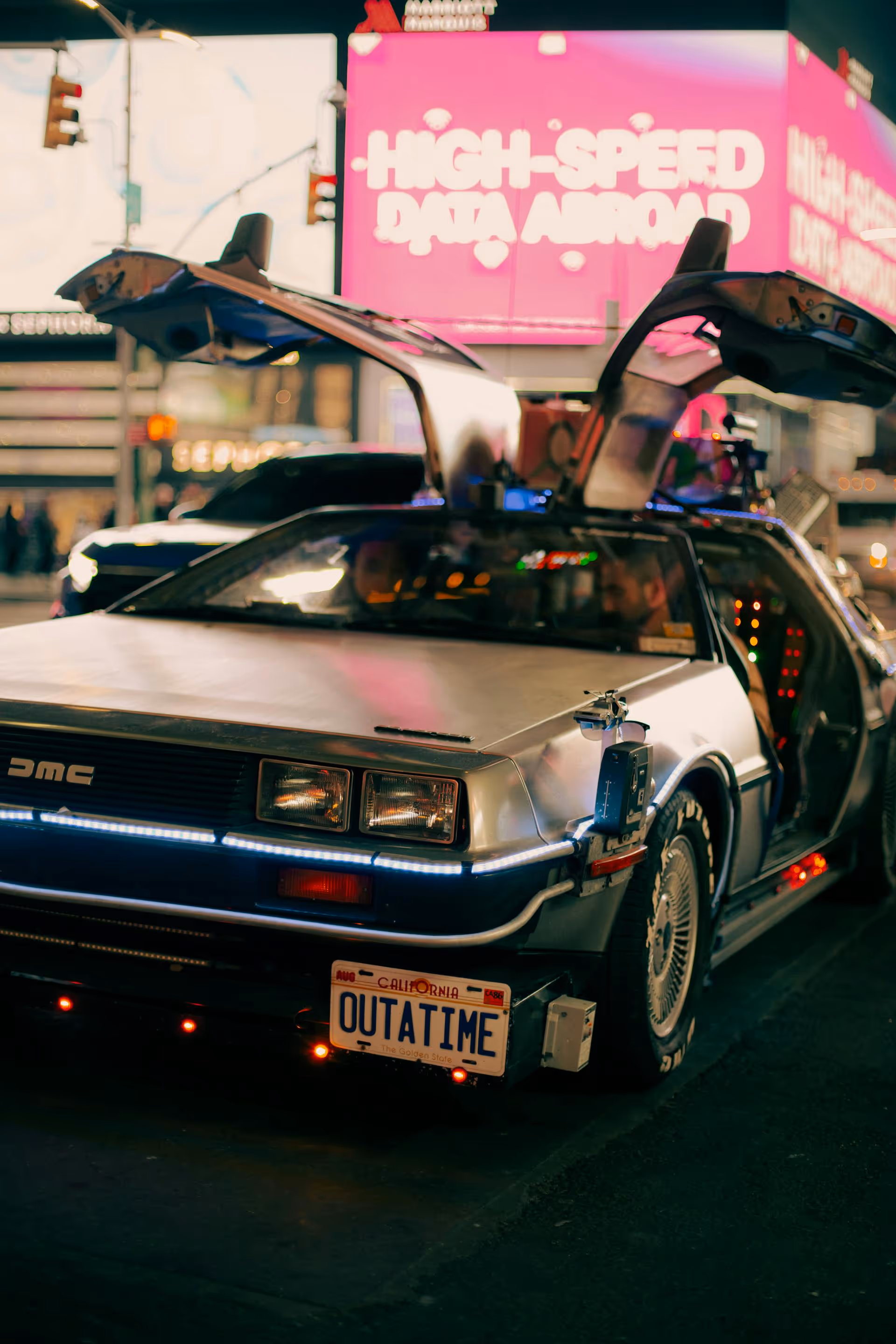 DeLorean sports car with gull-wing doors open and a California license plate reading OUTATIME, parked on a city street at night.