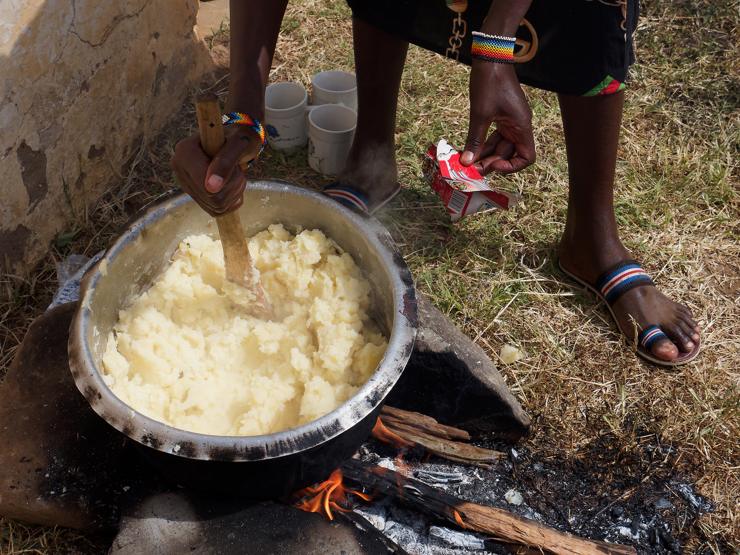 A woman stirs a large pot of ugly and adds salt over an open fire.