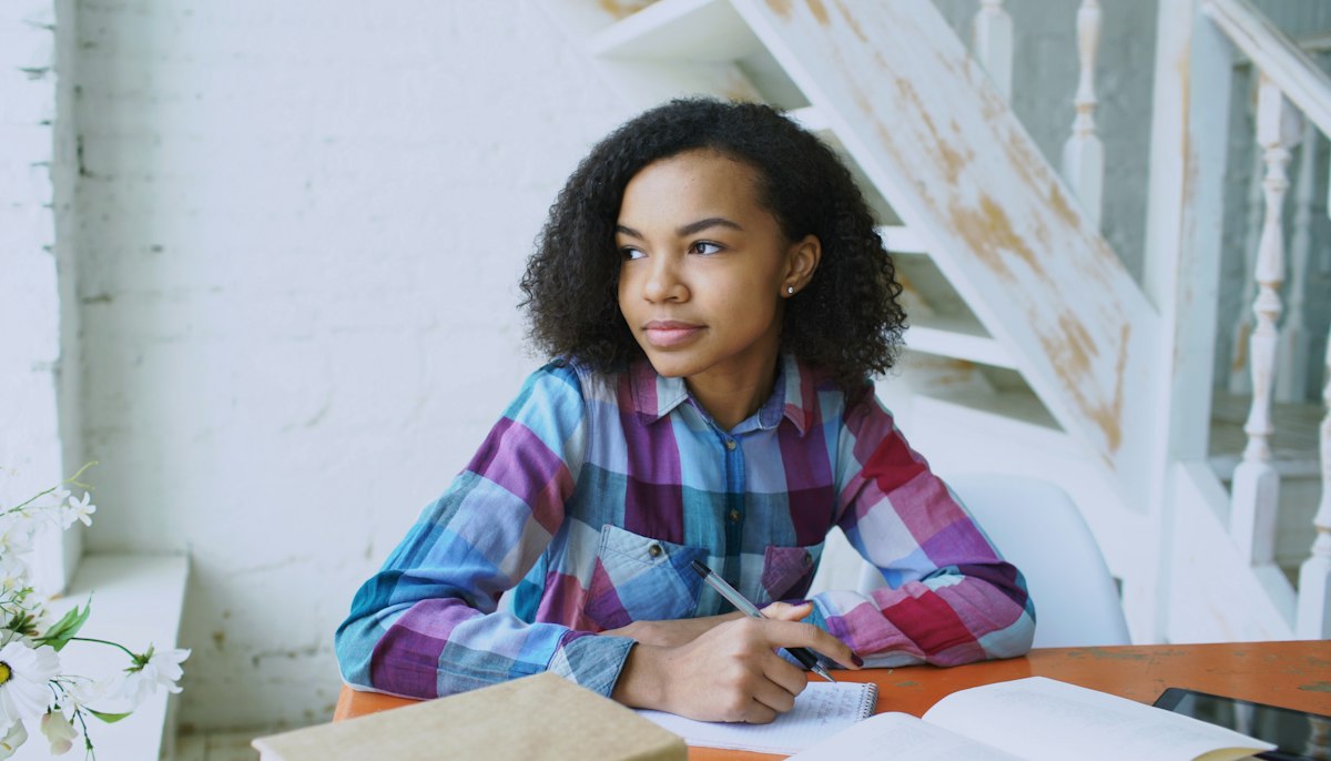 A young woman studying at a table with a notebook, looking out a window