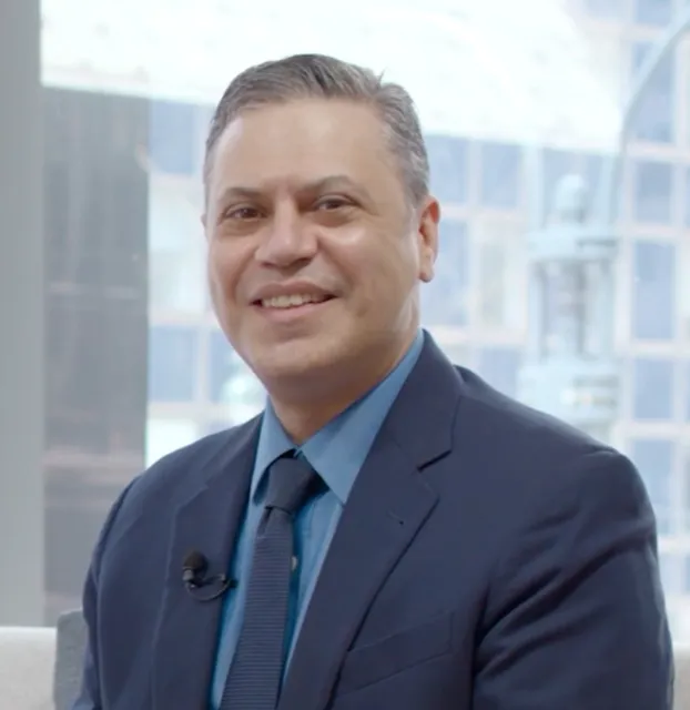 A profile photo of Dr. Bryan Wolynski wearing a blue suit and tie with a microphone clipped to his lapel, in an indoor setting with blurred buildings in the background.