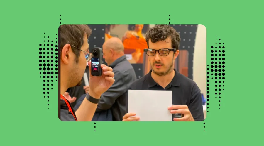 A man wearing the Envision Glasses, showing a document to another man who is recording on his phone