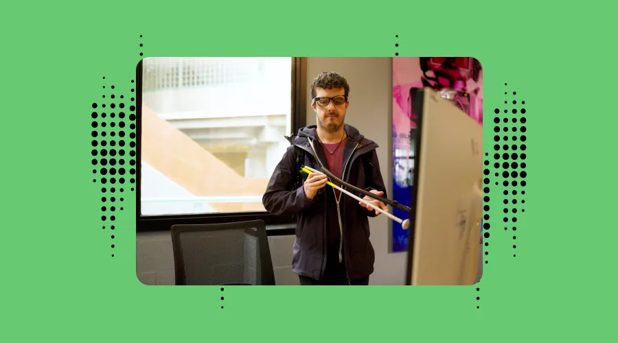 ben wearing envision glasses and folding his cane behind a desktop at work. the background is green with envision black dots