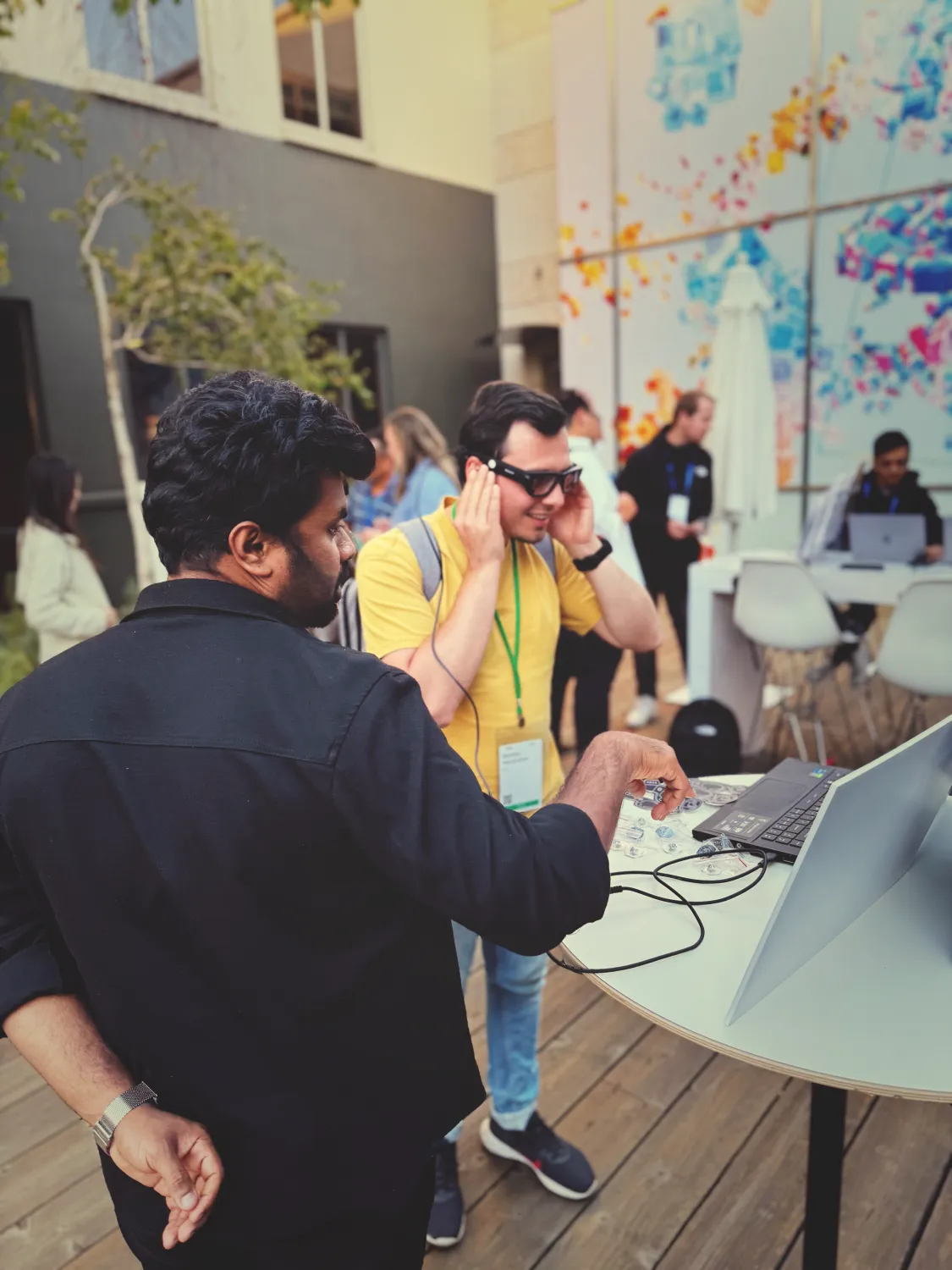 Alt: Me standing at a table with a laptop, demonstrating to another attendee wearing a yellow shirt and glasses our work with the Project Aria Glasses.