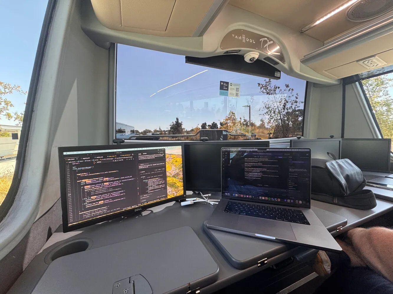 Alt: The interior of a van or mobile workspace, showing a desk setup with two computer screens. One screen displays code, while the other shows text. A laptop is also visible. The workspace is positioned in front of a large window offering a view of an outdoor area with trees and buildings in the distance. The van's interior features modern amenities and appears to be a custom mobile office setup.