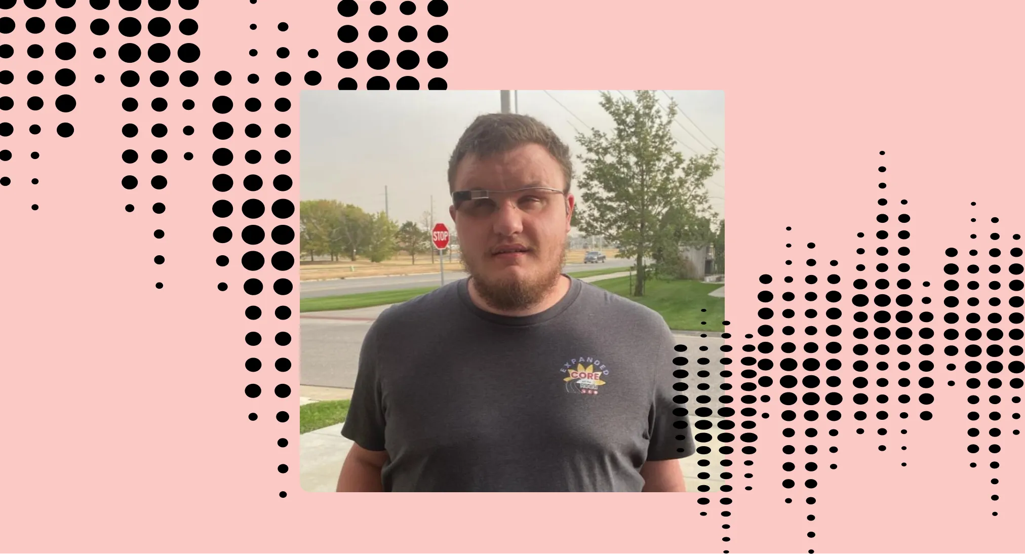 Jimmy Lewis is wearing the Envision Glasses wearing a black T-Shirt, and has some roads and trees as his background with a 'Stop' sign from the back of his right side.