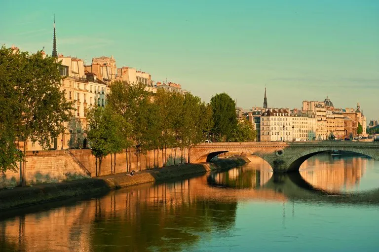 A view of the Seine River in Paris at sunset, with a charming bridge, tree-lined banks, and historic buildings reflecting in the calm water