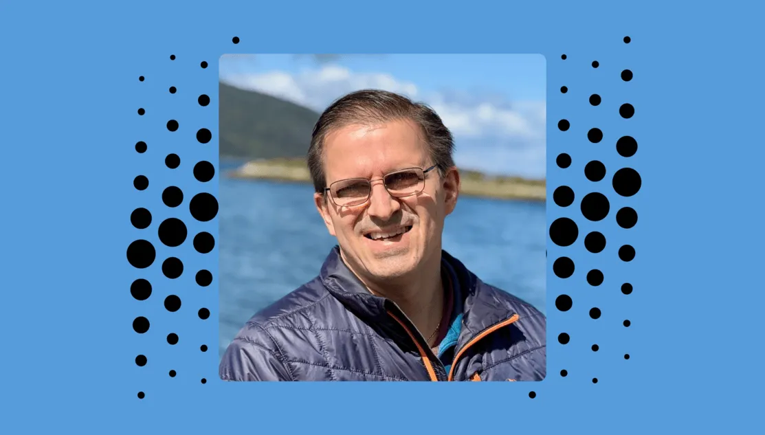 A picture within a blue frame is Jay, wearing glasses, smiling to the camera. He is wearing a black or dark blue winter jacket with orange zip. Behind him is a sea or a big river.