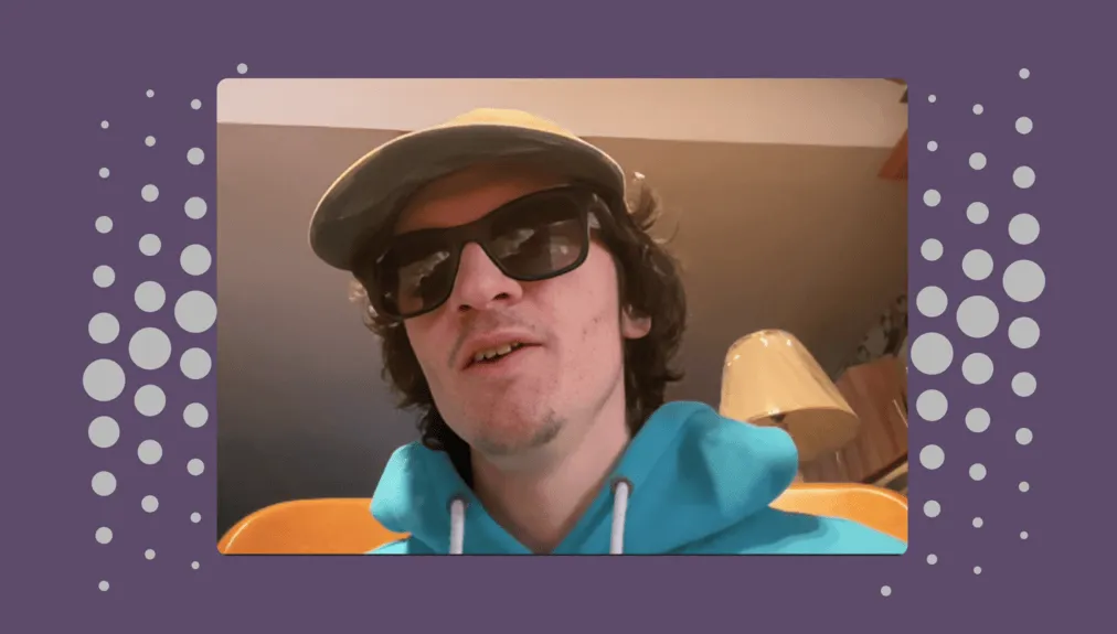 Scott wearing black sunglasses, beige baseball cap, and a turqiose hoodie. He seems to be sitting at home. Behind hime is a beige framed desk lamp.