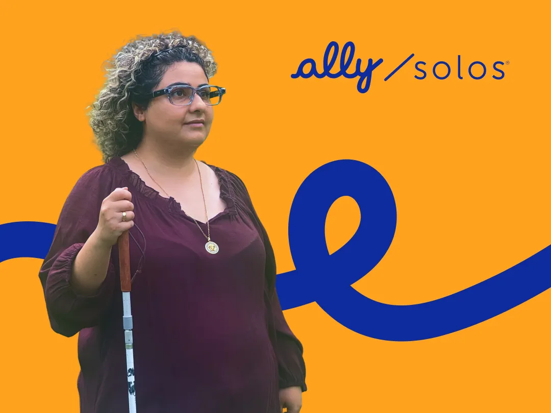 A woman with curly hair wearing smart glasses and a burgundy blouse stands holding a white cane. She has a gold pendant necklace and looks slightly to the side. The background is bright orange with a bold blue curved line and the ally / solos logo in the top right corner.