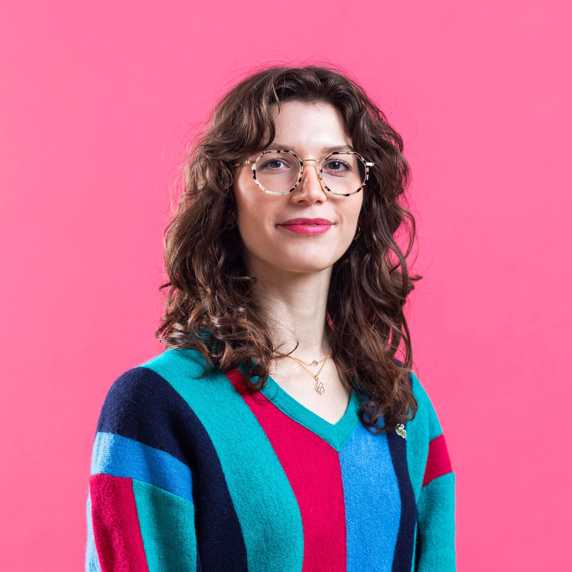 A headshot of Virginia wearing a colorful sweater on a pink background.