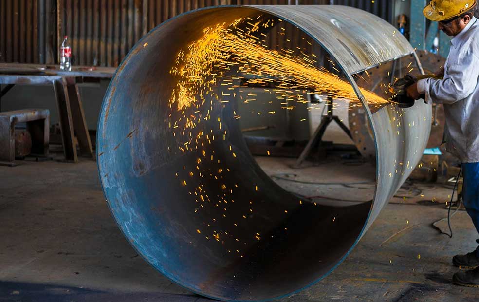 Worker wearing a yellow helmet grinding the edge of a large metal pipe, producing bright sparks in an industrial workshop.