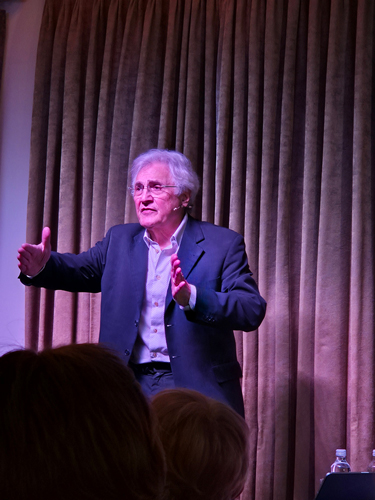 Older man with white hair and glasses speaking and gesturing on stage in front of closed brown curtains.