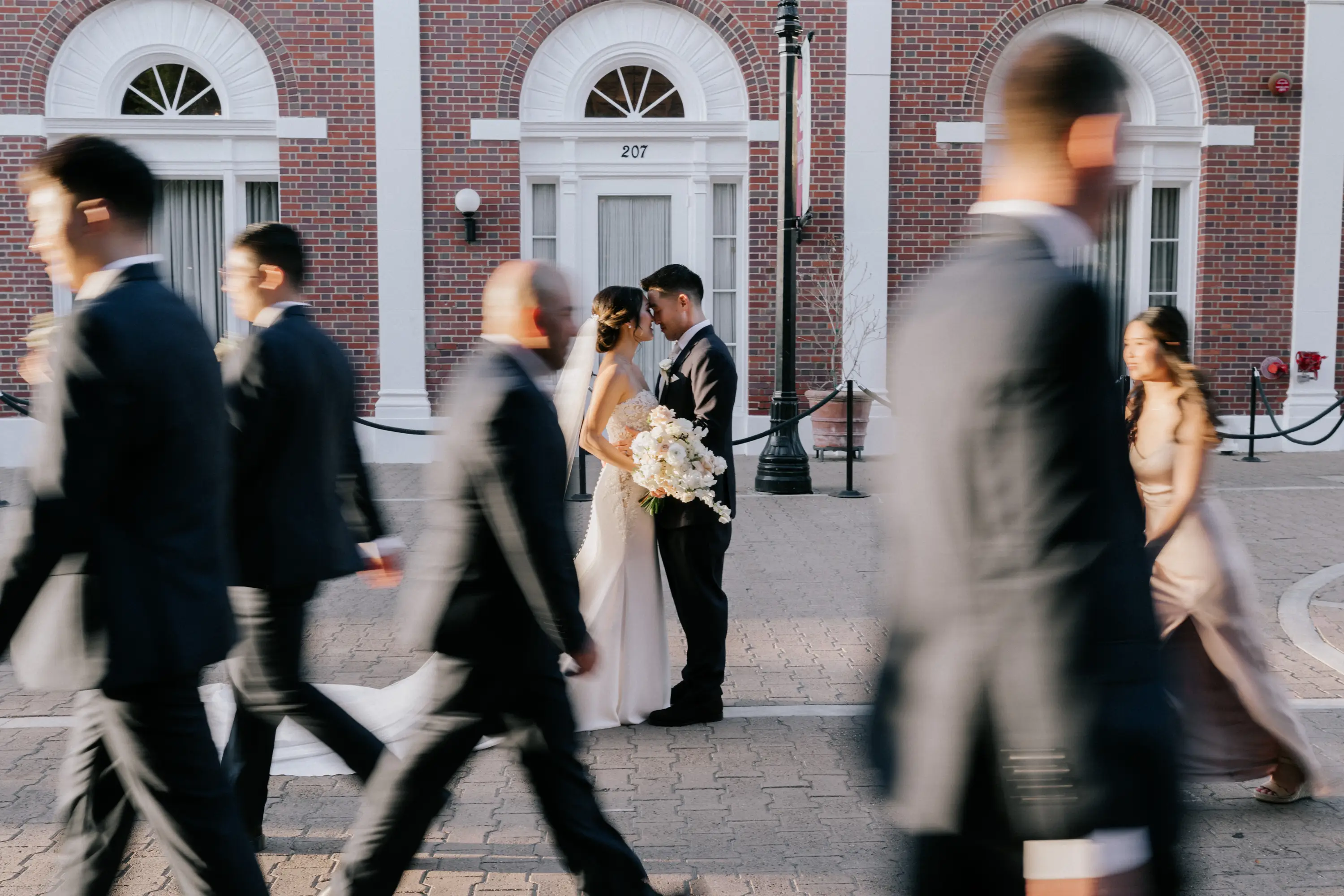 Bride and groom standing closely with foreheads touching while people in formal wear walk past them in blurred motion outside a brick building.