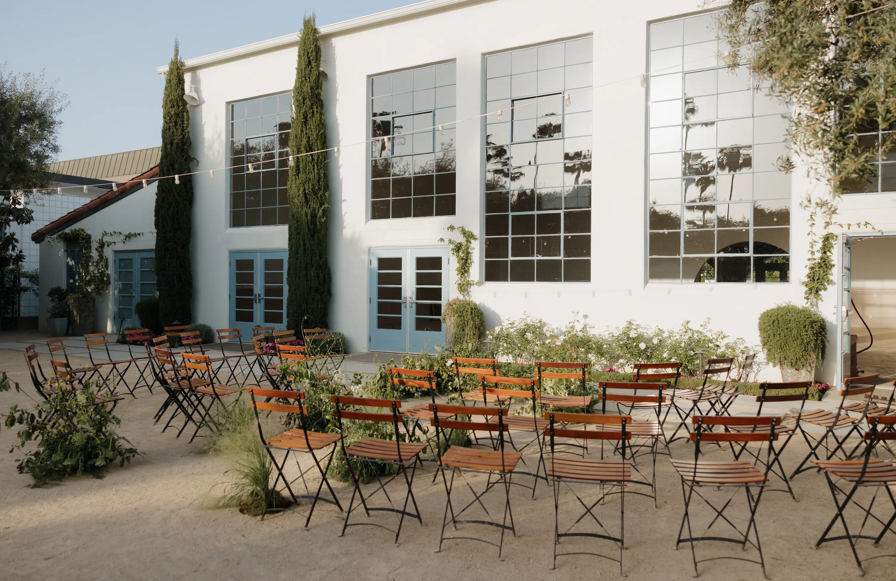 Outdoor seating area with wooden chairs arranged in rows in front of a white building with tall windows and blue doors, decorated with greenery and string lights.