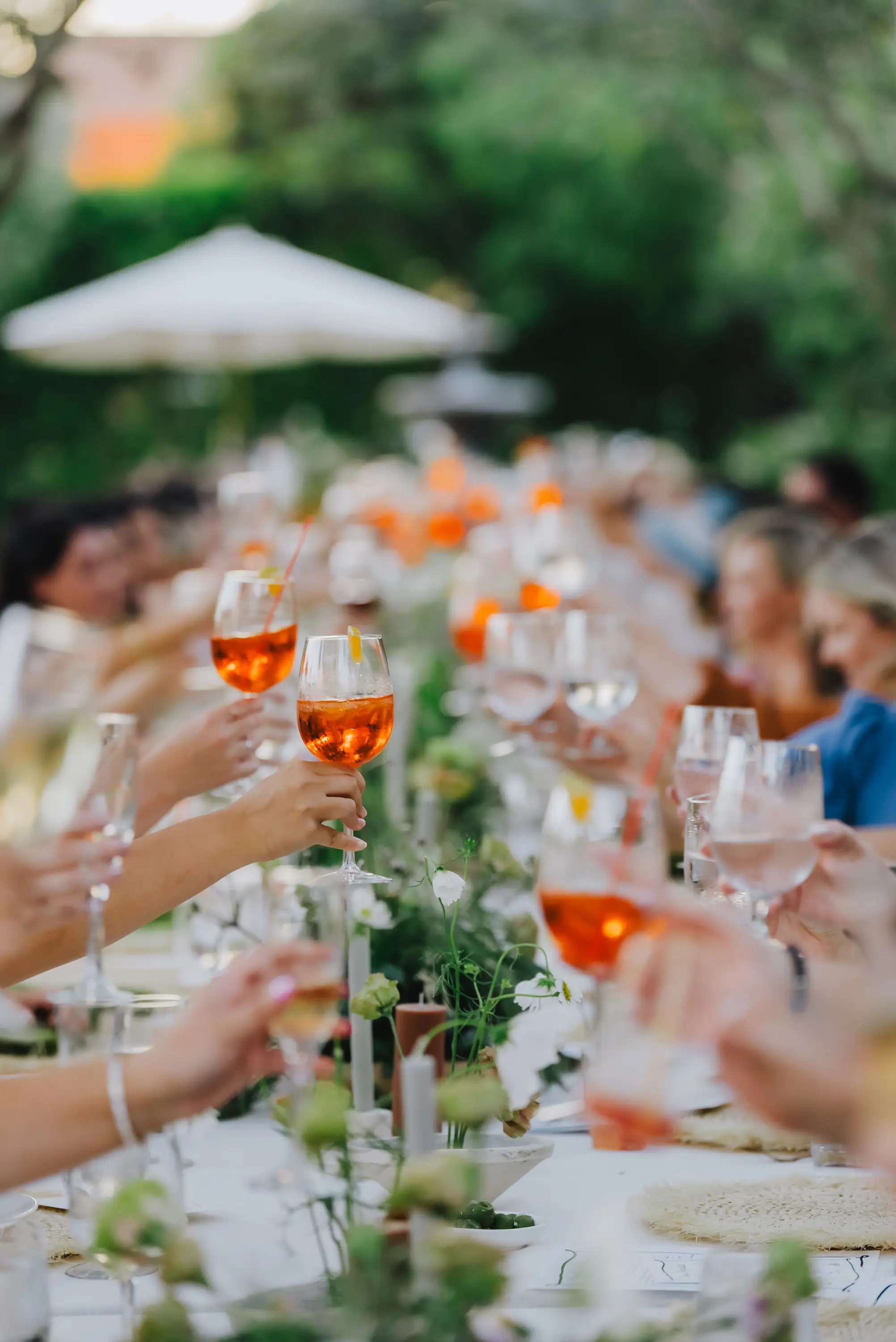 People raising glasses with orange cocktails in a toast at a long outdoor dining table decorated with flowers and candles.