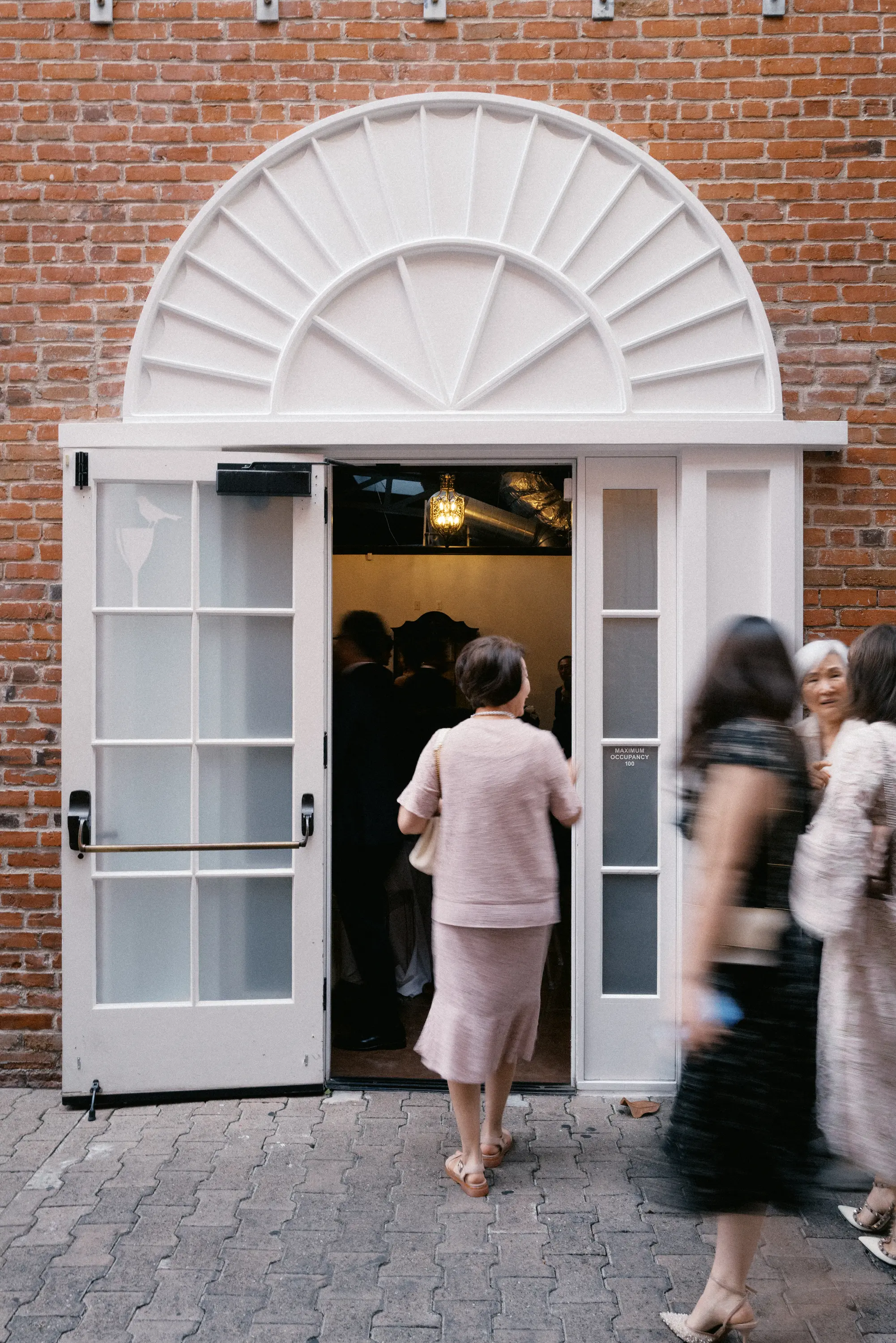 People entering and walking past an open white double door with frosted glass panels set in a red brick wall.