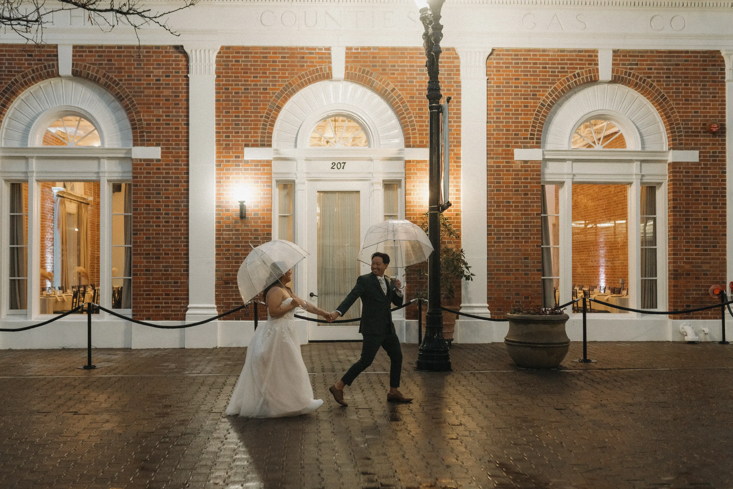 Bride and groom holding hands and walking with clear umbrellas on a wet brick pathway in front of an illuminated brick building at night.