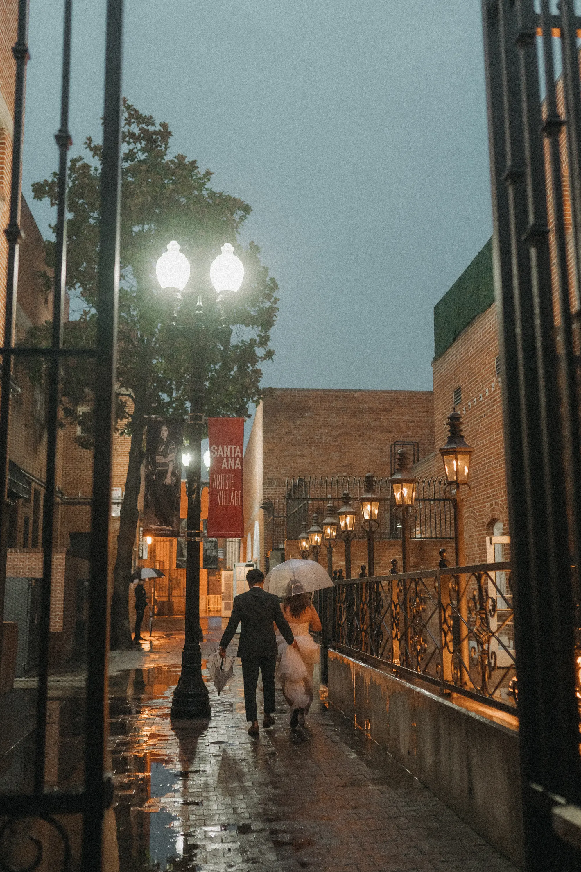 Couple dressed in formal attire walking under an umbrella along a wet street at dusk in Santa Ana Artists Village.