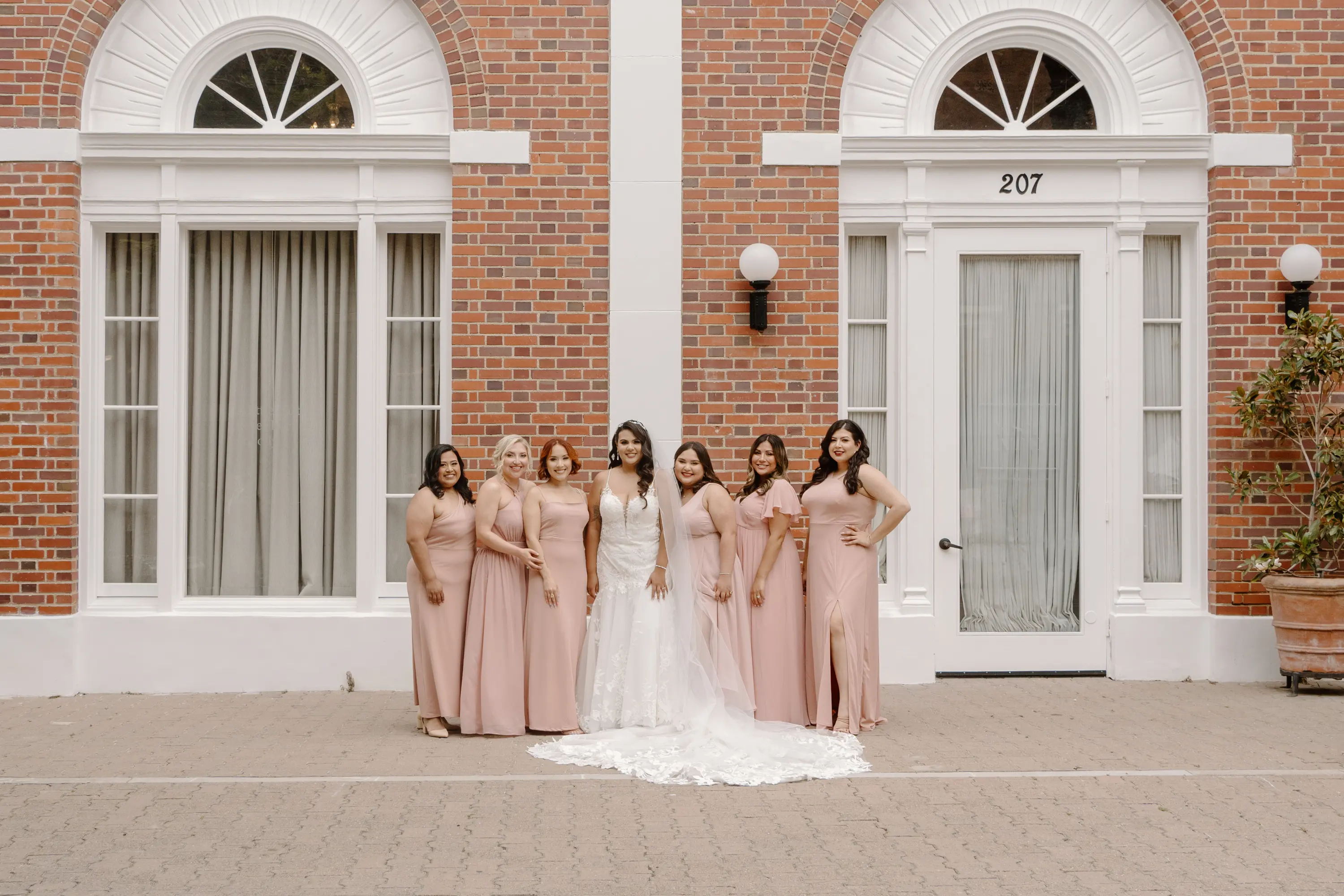 Bride in white wedding gown and six bridesmaids in blush pink dresses posing in front of a brick building with white-trimmed windows and door numbered 207.