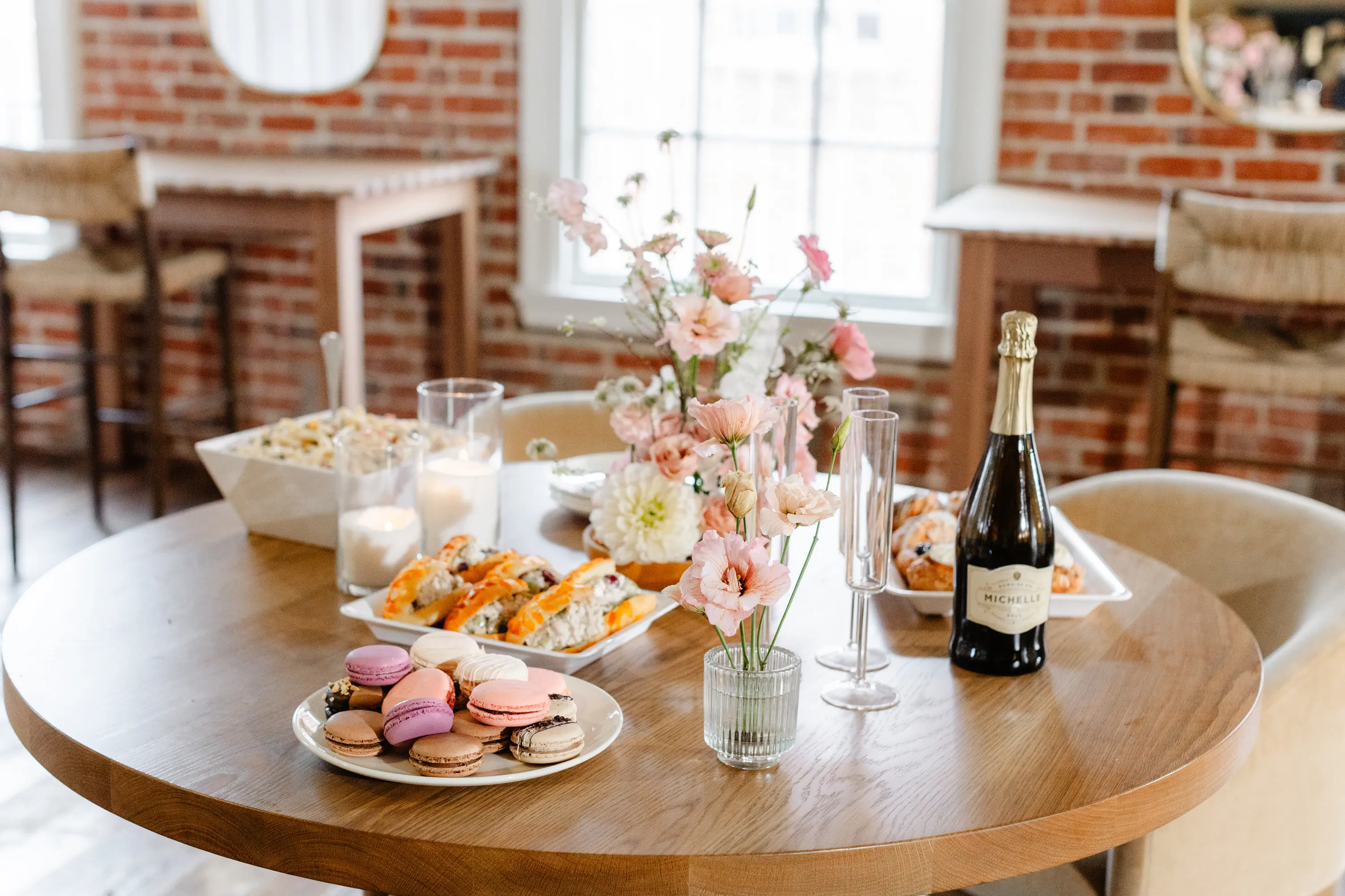 Round wooden table with macarons, pastries, a bottle of sparkling wine, two empty champagne flutes, lit candles, and pink floral arrangements in a bright room with brick walls.