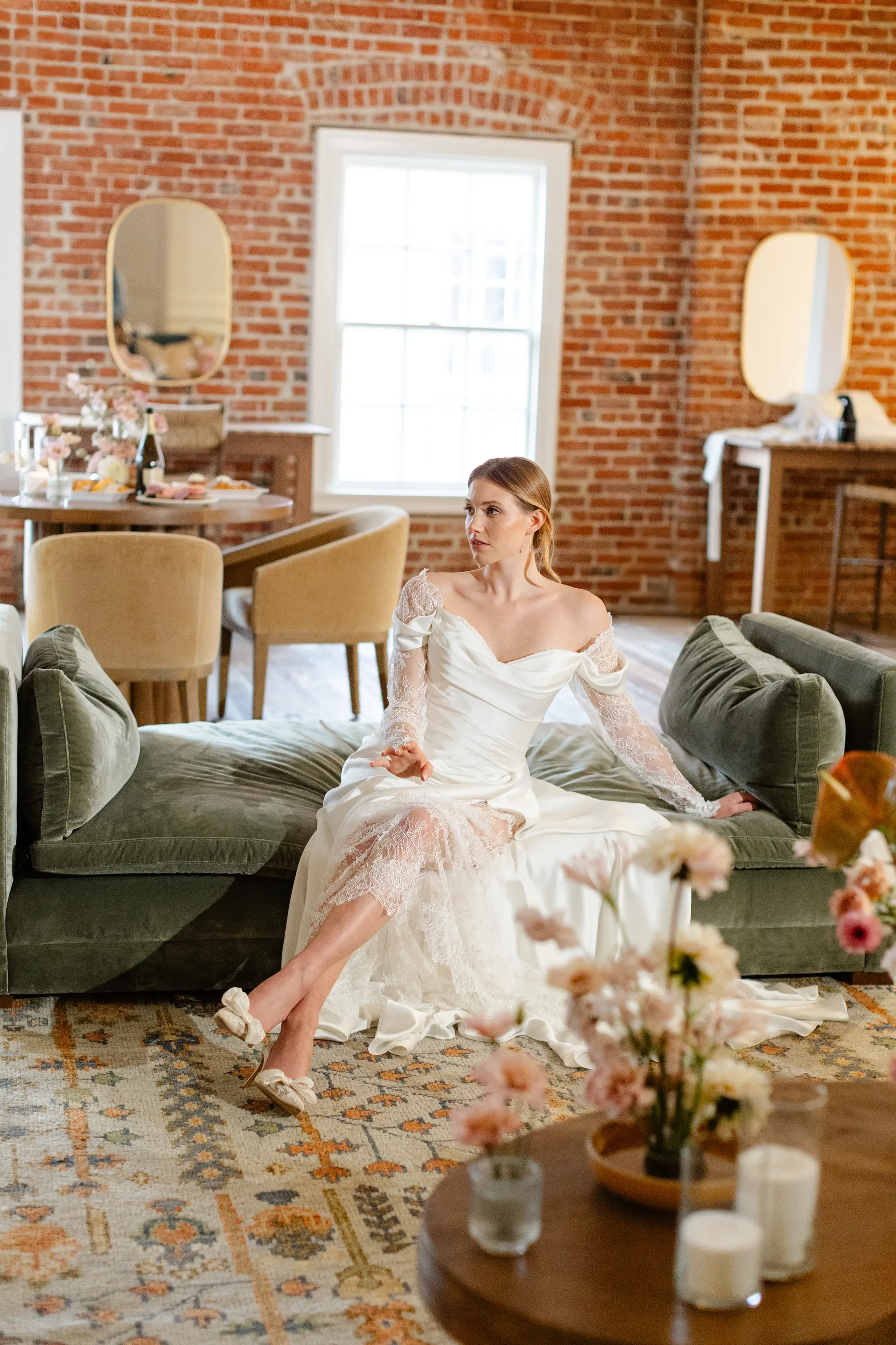 Woman in an off-shoulder white lace wedding dress sitting on a green sofa in a room with exposed brick walls and a large window.
