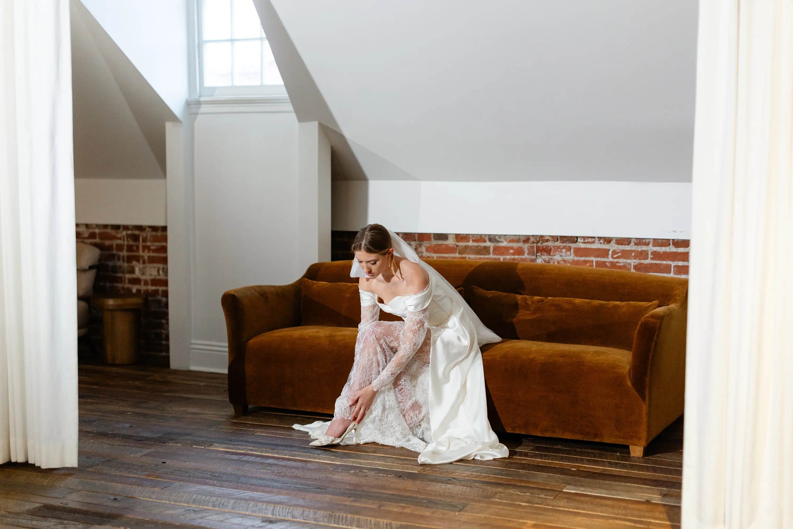 Bride in a white lace wedding gown sitting on a brown couch, putting on her shoe in a rustic room with wooden floors and brick walls.