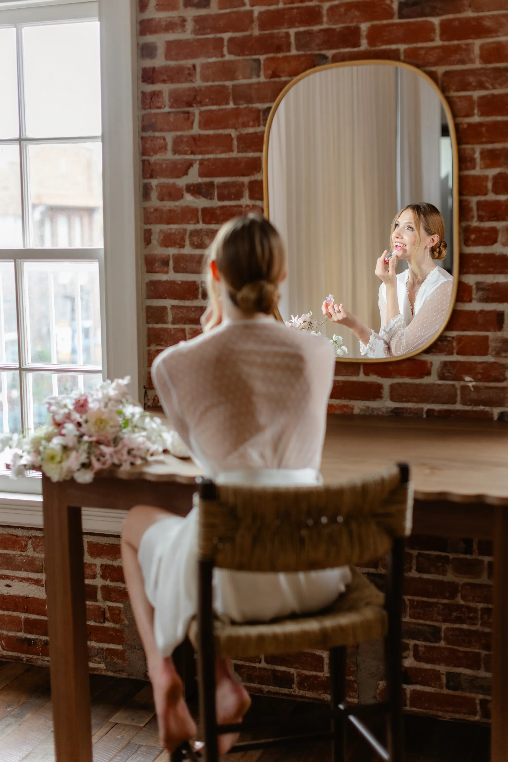 Woman in a white dress sitting on a chair applying lip balm in front of a mirror on a brick wall with a bouquet on the table.